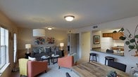 This single-floor residence offers a thoughtfully designed open-concept layout that seamlessly connects the living and kitchen areas. The living room is a welcoming and comfortable space, centered around a plush gray sofa and complemented by two rust-orange accent chairs that add a warm, vibrant touch. A round white coffee table sits at the heart of the seating arrangement, while colorful abstract wall art brings personality and energy to the neutral walls. Ceiling-mounted light fixtures provide consistent ambient illumination throughout the space. The kitchen is positioned adjacent to the living area, separated by a raised breakfast bar countertop with dark stone-like surfaces and bar-height stools — ideal for casual dining or entertaining. Cabinetry in warm wood tones lines the upper kitchen walls, adding a classic touch. A full-size refrigerator is visible, indicating a fully equipped kitchen space. Carpeted flooring flows through the living area, contributing to the cozy atmosphere, while the rustic wood-toned dining table in the foreground adds a natural, earthy contrast. A hallway visible from the living room leads to additional rooms, suggesting a functional floor plan designed for comfortable everyday living.