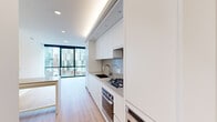 This thoughtfully designed one-bedroom, one-bathroom residence offers a clean, modern aesthetic across its well-appointed single floor. Light hardwood flooring flows throughout, complementing the bright, white palette that defines the interior.

The combined kitchen and living area serves as the heart of the home. The galley-style kitchen is outfitted with flat-front white cabinetry extending to the ceiling, a gas cooktop, stainless steel oven, integrated appliances, and a sleek undermount sink with a contemporary faucet. Recessed and cove lighting adds warmth overhead, while a neutral-toned island provides additional prep and dining space.

The generously sized bedroom features expansive floor-to-ceiling windows framed in bold black metal, offering sweeping views of the surrounding urban cityscape and filling the room with abundant natural light. A dedicated closet provides practical storage.

The bathroom is finished in a refined, neutral palette with large-format floor tiles, full-height grey wall tiles, a frameless glass-enclosed shower, a floating white vanity with an undermount sink, and a wide mirror. A hallway connects all rooms efficiently, completing the functional layout of this well-designed urban residence.