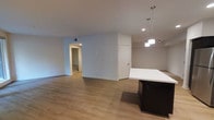 This single-floor space showcases a clean, modern design with an open-concept layout that seamlessly connects the kitchen and living areas. The kitchen features a sleek island with a crisp white countertop and dark base cabinetry, illuminated by contemporary pendant lights that add a stylish focal point to the space. Recessed lighting throughout ensures consistent, practical illumination across the entire area.

The flooring consists of warm-toned hardwood-style planks that run continuously throughout, lending a cohesive and inviting feel to the space. Neutral gray-white walls provide a versatile backdrop that complements the modern aesthetic. A hallway visible in the background leads to additional rooms, suggesting a well-organized floor plan with clearly defined private and communal zones. A white interior door provides access to a separate room or utility area, adding to the functional layout.

The open layout creates a generous sense of space, making the area feel bright and airy. The combination of recessed lighting, pendant fixtures, and neutral tones contributes to a contemporary ambiance that is both practical and visually appealing.