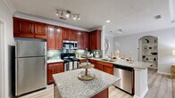 Located at 5375 Sugarloaf Pkwy in Lawrenceville, GA, this well-appointed two-bedroom, two-bathroom residence offers comfortable single-floor living across a thoughtfully designed layout. The kitchen is a standout feature, showcasing rich cherry wood cabinetry, granite countertops, a subway tile backsplash, stainless steel appliances, and a center island with additional counter space — all set atop light hardwood-style flooring. The kitchen flows seamlessly into the living room, which is distinguished by an elegant tray ceiling with crown molding and large French doors that invite abundant natural light and outdoor views. Both bathrooms are well-finished with granite vanity tops, tile-accented tub-shower combinations, and hardwood-style flooring. The spacious primary bedroom features plush carpeting, a ceiling fan, and generous natural light, while the second bedroom offers a similarly comfortable setup. Two closets and two hallways provide ample storage and circulation throughout the unit. A dedicated laundry area with in-unit washer hookups adds everyday convenience. Neutral gray tones and consistent finishes throughout lend the space a cohesive, modern aesthetic suitable for a variety of lifestyles.