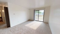 This single-floor unit offers a practical and comfortable layout with a clean, neutral aesthetic throughout. The living area is generously proportioned, featuring wall-to-wall carpeting, bright white walls, and a ceiling fan that promotes airflow and comfort. A sliding glass door fills the space with natural light and opens directly onto a private wooden balcony, which overlooks a landscaped exterior with views of the surrounding neighborhood and parking area. The balcony features a freshly built wood railing, providing an outdoor retreat.

A second carpeted room, suitable as a bedroom or flex space, includes a sliding window that frames views of nearby trees, allowing natural light to filter in. The kitchen is efficiently designed with warm wood-toned cabinetry, white countertops, a double sink, and white appliances including a refrigerator, electric range, and dishwasher. Wood-look flooring adds durability to this functional space.

The bathroom is well-appointed with a wide vanity featuring a large mirror and integrated lighting, a white tub-shower combo with tiled surround, and wood-look flooring that ties in with the kitchen. The overall ambiance is clean, simple, and move-in ready.