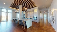 Located at 4011 Tryon Park Rd in Charlotte, NC, this thoughtfully designed single-floor space blends modern aesthetics with warm, inviting interiors. Rich hardwood flooring flows throughout, unifying the open-plan layout and lending a sense of continuity across each area.

The kitchen is a standout centerpiece, featuring an expansive waterfall-edge island with seating for multiple guests. Striking slatted wood ceiling details overhead are complemented by contemporary chandelier lighting, while a textured stone backsplash and built-in stainless steel appliances add both function and visual interest.

Adjacent lounge areas are thoughtfully appointed with layered seating arrangements, including a plush teal velvet sofa, a sculptural marble coffee table, and bold curated artwork that energizes the walls. Tray ceilings with accent lighting — finished in deep teal and charcoal tones — add dramatic architectural depth and define each social zone within the open floor plan.

Floor-to-ceiling windows flood the space with abundant natural light and offer open views of the surrounding landscape, while sheer drapery softens the brightness and creates an airy atmosphere. A media wall with built-in cabinetry and a large-format display anchors a dedicated entertainment area, rounding out this well-appointed, design-forward space.