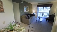 This single-floor space offers a comfortable and functional layout that seamlessly connects the dining and living areas. The living room is anchored by a plush, neutral-toned sofa accented with dark decorative pillows, complemented by a glass-top coffee table with an ornate metal base that adds an elegant touch. A warm table lamp provides ambient lighting alongside a framed piece of wall art displayed on a striking sage-green accent wall, which contrasts tastefully against the surrounding neutral beige tones. Soft wall-to-wall carpeting runs throughout, contributing to a cozy atmosphere underfoot. A sliding glass door floods the space with natural light and opens to a private balcony, offering views of mature trees and greenery beyond. The dining area, visible in the foreground, features a glass-top table set with coordinated dinnerware and green cloth napkins, creating an inviting setting for meals. A valance in deep burgundy frames the large window, adding a refined decorative detail. The overall ambiance is warm, understated, and well-appointed, making practical use of the available space while maintaining a welcoming aesthetic.
