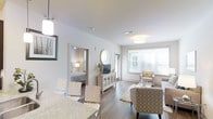 This single-floor residence offers a welcoming and cohesive open-concept design that connects the living, dining, and bedroom areas in a smooth, functional flow. The living area is bathed in natural light streaming through large windows fitted with blinds, creating a bright and airy atmosphere. Smooth white ceilings with flush-mount and pendant lighting fixtures provide clean, even illumination throughout the space. Rich hardwood-style flooring runs consistently across the main living area, adding warmth and a sense of continuity. The living space is thoughtfully arranged with patterned upholstered seating, a soft area rug, and a glass-top coffee table, all contributing to a refined yet comfortable ambiance. A media console with a circular mirror above it serves as a stylish focal point along the main wall. The dining area features a granite-topped counter with modern seating, blending practicality with contemporary aesthetics. An open doorway leads directly into the bedroom, which is softly lit and furnished with a classic bed and lamp, maintaining the home's consistent neutral palette. Overall, the space presents a clean, modern aesthetic with thoughtful finishes and a light, inviting atmosphere throughout.