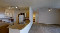 This single-story residence in Waco, Texas offers an open-concept layout that seamlessly connects the kitchen and living areas. The kitchen is equipped with warm-toned cabinetry, including upper cabinets with decorative glass-front detail, and features a peninsula with a built-in sink and extended countertop that doubles as a casual dining or workspace surface. A full-size refrigerator anchors the kitchen, and track lighting overhead provides direct, functional illumination. The flooring transitions from the kitchen area into carpeted living space, adding a sense of comfort and warmth to the main living zone. A partial wall divides the kitchen from the rest of the space while maintaining an open feel throughout. Neutral white walls and standard ceiling height keep the interior feeling clean and cohesive. A doorway visible from the main living area leads to an additional room, suggesting a bedroom or utility space beyond. Ceiling vents throughout indicate a central HVAC system. The overall layout is practical and straightforward, making efficient use of the available footprint.