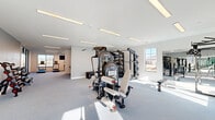 Located at 20021 Chad Hittle Dr in Westfield, Indiana, this single-floor fitness facility offers a thoughtfully designed space built for serious training. The expansive exercise room is the centerpiece, bathed in natural light from multiple windows and illuminated by sleek recessed linear lighting along bright white ceilings. The open floor plan provides ample room for a wide range of equipment, including a multi-station cable and weight machine, a treadmill, stair climber, elliptical, rowing machine, and a dedicated free-weight area with a squat rack. Full-length wall mirrors along one side enhance the sense of space and support form-conscious training. A bold steel-framed glass wall and door partition elegantly separates sections of the facility, adding a modern architectural touch. The hallway features a striking wood-panel accent wall with a dual drinking fountain station, complemented by a warm reception area. The ADA-accessible bathroom continues the wood-tile design aesthetic with horizontal slat-style wall tile, stainless steel fixtures, and durable floor tile. Throughout, the clean white palette, neutral carpet flooring, and modern finishes create a bright, professional, and welcoming fitness environment.