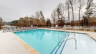 Located at 13100 Margie Ln in Raleigh, NC, this outdoor amenity space serves as a community gathering area centered around a large, sparkling swimming pool. The pool is finished with light stone coping and is surrounded by a generous concrete deck lined with multiple lounge chairs, offering ample space for relaxation and recreation. A covered patio structure with white columns and a pitched roof provides a shaded retreat directly adjacent to the pool, making it an ideal spot for outdoor gatherings. The entire pool area is enclosed by a black wrought iron fence, providing both security and a clean, cohesive aesthetic. Manicured landscaping with mulched beds and low shrubs frames the perimeter, adding a polished finish to the grounds. Stone retaining walls with iron railings and concrete steps offer structured access points throughout the property. The surrounding multi-story residential buildings feature a mix of brick and siding exteriors with balconies overlooking the amenity area. A concrete walkway runs along the perimeter of the buildings, connecting the various sections of the community. Mature trees visible beyond the fencing contribute a natural backdrop, enhancing the overall outdoor ambiance of this well-appointed community amenity space in Raleigh, North Carolina.