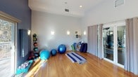 This single-level yoga and fitness studio is thoughtfully designed to support a wide range of movement and wellness activities. The studio features warm hardwood flooring throughout, providing a durable and attractive surface ideal for yoga, stretching, and light exercise. Soaring ceilings amplify the sense of openness, while recessed lighting and wall-mounted sconces create a balanced, inviting atmosphere.

A large floor-to-ceiling mirror spans one wall, allowing practitioners to monitor their form and enhancing the perception of space. The accent wall is finished in a deep navy blue, adding a calm, focused energy to the room. A sliding glass door floods the studio with natural light and opens directly to an outdoor balcony, offering pleasant views of the surrounding landscaped grounds.

The studio is well-stocked with equipment, including yoga mats, stability balls in multiple sizes, foam blocks, dumbbells, step platforms, and a medicine ball rack. Curtained glass doors connect the space to an adjacent fitness room equipped with cardio machines, providing convenient access to additional workout options. Overall, the studio offers a serene yet functional environment suited for yoga, pilates, and general fitness training.