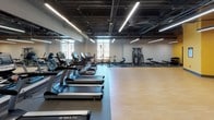 Located at 175 Market St in Westlake, Ohio, this single-floor fitness facility offers a thoughtfully designed workout environment suitable for a range of exercise routines. The main cardio area is expansive and open, housing a row of TRUE-brand treadmills, elliptical machines, stationary bikes, and a rowing machine. Large windows along the perimeter allow natural light to fill the space, while linear LED lighting runs across the exposed ceiling, which features visible ductwork lending an industrial aesthetic. Full-length mirrors along one wall amplify the sense of space and support proper form during workouts.

A dedicated strength and functional training zone is equipped with cable machines, resistance equipment, and a water cooler station with paper towel dispenser for convenience. The flooring transitions between rubber sport flooring in the equipment zones and smooth hardwood in the open exercise areas.

A separate studio room features vibrant yellow accent walls, hardwood flooring, a wall-mounted display screen, punching bags, medicine balls, stability balls, resistance bands, and foam rollers organized on a multi-tier rack. This versatile studio is well-suited for group classes, boxing training, or functional fitness sessions.