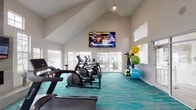 Located at 7938 Island Club Drive in Indianapolis, Indiana, this single-floor fitness center is a well-appointed community amenity designed to support a comprehensive workout experience. The space is defined by dramatic vaulted ceilings that soar to impressive heights, creating an open and airy atmosphere throughout. A striking circular clerestory window crowns one end of the room, flooding the interior with natural light alongside multiple large double-hung windows that frame views of the surrounding community.

The main workout area is outfitted with a full range of cardio equipment, including treadmills, ellipticals, and an upright bike, all positioned on vibrant teal-toned carpet tiles that lend a modern, energetic feel to the space. Spin bikes, a dumbbell rack, a weight bench, and a medicine ball storage tower round out the strength and cross-training offerings. A large wall-mounted television keeps members entertained during workouts, while built-in ceiling speakers provide ambient sound.

A sleek, tiled accent wall houses a linear electric fireplace beneath another mounted television, serving as a stylish focal point. An adjacent alcove features built-in cabinetry and countertops, offering a convenient refreshment or resource station. Sliding glass doors open to an outdoor deck with seating, providing a refreshing connection to the outdoors. Wall mirrors enhance the sense of space and support proper form during exercise sessions.