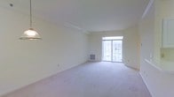 This single-floor residential space offers a clean, understated aesthetic defined by neutral tones and an open, airy layout. The main living area is generously proportioned, featuring soft wall-to-wall carpeting in a light beige hue that complements the crisp white walls and ceiling throughout. Crown molding adds a subtle architectural detail, lending a polished finish to the space. A large sliding glass door serves as a focal point, flooding the interior with natural light and providing direct access to an outdoor balcony or patio area, where glimpses of greenery can be seen beyond. A wall-mounted heating unit is conveniently positioned near the exterior wall, ensuring year-round comfort. The open floor plan creates a versatile living environment, easily adaptable to a variety of furniture arrangements and lifestyle needs. The overall ambiance is calm and inviting, with the bright, light-filled interior creating a sense of spaciousness. The minimalist design palette allows the architectural elements — from the smooth ceilings to the well-proportioned room dimensions — to take center stage, making this a welcoming and functional living space.