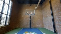This amenity suite offers residents a well-rounded collection of shared spaces designed for recreation, fitness, and community engagement. The standout feature is the indoor basketball court, an impressive space defined by its exposed brick walls that rise dramatically to a tall ceiling with visible industrial ductwork overhead. A regulation-height glass backboard and hoop are anchored at center court, while the playing surface features a tiled sport floor with a custom logo at mid-court, surrounded by green boundary markings. The raw, industrial character of the brick enclosure lends the court an energetic and urban aesthetic rarely found in residential amenity spaces. Beyond the basketball court, the suite includes a fitness center equipped for residents seeking convenient on-site exercise options. A media room provides a dedicated space for entertainment and group screenings, offering a comfortable retreat for relaxation. The community room rounds out the offerings, serving as a versatile gathering space for social events and resident activities. Together, these amenities form a comprehensive recreational hub that caters to a variety of lifestyles and interests within a single, thoughtfully designed floor.