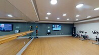 Located at 1300 Tribute Center Dr in Raleigh, NC, this single-floor amenity space combines fitness and recreation into a cohesive, well-designed facility. The exercise studio is outfitted with resilient bamboo-style flooring, a full-length wall mirror with a ballet barre, and recessed circular ceiling lights set within a tray ceiling. The space supports a range of activities, including barre workouts, stretching, and group fitness classes, with a wall-mounted flat-screen TV and a control panel for interactive programming. Spin cycles, medicine balls on a rack, and a standing punching bag round out the fitness offerings. An adjacent area houses additional spin cycles alongside racks of kettlebells, dumbbells, stability balls, and step platforms, providing a comprehensive strength and cardio training environment. The games room transitions seamlessly from the fitness areas and features dark patterned carpet, tray ceilings with recessed and track lighting, and multiple wall-mounted flat-screen TVs. A full-size pool table anchors the room, complemented by a shuffleboard table and high-top bar seating with bistro-style chairs, creating an inviting social atmosphere. The overall design balances functionality and comfort, making this facility suitable for both active workouts and casual leisure activities.