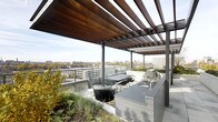 This rooftop terrace is a thoughtfully designed outdoor living space that combines modern architectural elements with functional amenities and sweeping urban views. A striking pergola constructed from rich warm-toned wood slats supported by sleek steel posts defines the covered zone, providing partial shade while maintaining an open, airy feel and framing the expansive skyline beyond.

Beneath the pergola, a dining area accommodates several guests around a rectangular table paired with contemporary wire chairs, making it well-suited for outdoor entertaining. Adjacent to the seating area, a built-in outdoor kitchen features a stainless-style countertop with an integrated sink and grill station, offering everything needed for al fresco cooking and hosting.

Integrated planter beds line portions of the terrace perimeter, filled with established greenery and ornamental grasses that soften the urban hardscape and add a naturalistic quality to the space. A low glass-panel railing runs along the terrace edge, preserving unobstructed panoramic views of the surrounding cityscape, including treetops, rooftops, and open sky. The overall design balances industrial materials with organic textures, creating a refined and versatile outdoor retreat.