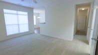 This single-floor space is designed with an open-concept layout that seamlessly connects the main living area to the dining space beyond. The living area features neutral-toned wall-to-wall carpeting that extends throughout, lending a clean and comfortable feel underfoot. Crisp white walls and clean baseboards contribute to a bright, minimalist aesthetic throughout the space.

A ceiling fan is installed overhead in the living area, providing both functionality and a subtle design element. Natural light filters into the room through a large window along the left wall, brightening the interior and enhancing the airy, open atmosphere.

Transitioning toward the rear of the space, a partial wall divides the living area from the dining space, which features contrasting hard-surface flooring. An elegant chandelier hangs above the dining area, offering a more refined lighting fixture that complements the overall neutral palette. The raised countertop or bar-height surface visible in the transition zone suggests a kitchen area is nearby, making this an efficient and well-connected floor plan ideal for everyday living and entertaining.