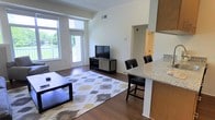 This single-floor space offers a thoughtfully designed open-concept layout that seamlessly connects the living and kitchen areas, creating a cohesive and functional environment. Rich, dark hardwood flooring extends throughout, anchoring the space with warmth and elegance.

The living area is bright and welcoming, benefiting from large floor-to-ceiling windows and a glass door that flood the room with natural light and provide views of the surrounding greenery. The generous proportions of the room allow for comfortable furniture arrangements, as evidenced by the spacious seating area complemented by a patterned area rug.

The kitchen features a raised granite-topped breakfast bar that serves as a natural divider between the living and cooking spaces. Warm wood cabinetry adds a contemporary touch, while bar-height seating along the counter makes it ideal for casual dining. The kitchen opens conveniently toward a hallway leading to additional areas of the unit.

Clean white walls and a neutral color palette throughout the space enhance the sense of airiness and light. The combination of modern finishes, natural light, and an open floor plan makes this a versatile and inviting space well-suited for both relaxing and entertaining.