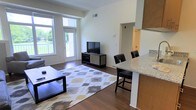 This single-floor space offers a thoughtfully designed open-concept layout that seamlessly connects the living and kitchen areas, creating a cohesive and functional environment. Rich, dark hardwood flooring extends throughout, anchoring the space with warmth and elegance.

The living area is bright and welcoming, benefiting from large floor-to-ceiling windows and a glass door that flood the room with natural light and provide views of the surrounding greenery. The generous proportions of the room allow for comfortable furniture arrangements, as evidenced by the spacious seating area complemented by a patterned area rug.

The kitchen features a raised granite-topped breakfast bar that serves as a natural divider between the living and cooking spaces. Warm wood cabinetry adds a contemporary touch, while bar-height seating along the counter makes it ideal for casual dining. The kitchen opens conveniently toward a hallway leading to additional areas of the unit.

Clean white walls and a neutral color palette throughout the space enhance the sense of airiness and light. The combination of modern finishes, natural light, and an open floor plan makes this a versatile and inviting space well-suited for both relaxing and entertaining.