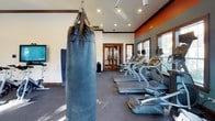This fitness center is thoughtfully designed to support a wide range of workout styles within a single, open-plan floor. The space is finished with a bold color palette of orange accent walls, teal-gray tones, and warm wood trim that frames the windows and doorways, lending the facility a distinctive, energetic character.

The cardio area houses a row of treadmills equipped with individual screens, positioned along windowed walls that allow ample natural light to fill the room. Adjacent to this is a dedicated spinning studio featuring multiple Spinning-brand bikes arranged in rows, complemented by a large wall-mounted flat-screen television and a floor-to-ceiling mirror that visually expands the space.

The strength training zone is anchored by a substantial multi-functional power rack offering cable attachments, TRX suspension straps, and storage for free weights and medicine balls. A heavy leather punching bag hangs from an overhead mount, supporting boxing and functional fitness training. Dumbbells are accessible along the perimeter.

Recessed ceiling lighting and a ceiling fan ensure comfort throughout workouts, while multiple windows provide views of the surrounding outdoor area, creating a bright and motivating environment overall.