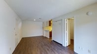 This single-floor space offers a clean, functional layout designed to maximize usability. The main living area is anchored by rich, dark hardwood-style flooring that extends throughout, providing a warm and cohesive aesthetic. White walls and a textured ceiling reflect light effectively, keeping the space feeling bright and open. A recessed light fixture adds focused illumination to the living area, complementing the natural simplicity of the room.

The kitchen is partially open to the living space, separated by a raised countertop peninsula that serves as both a prep surface and a casual dining or serving area. Warm wood-toned cabinetry lines the kitchen walls, offering practical storage, while upper white cabinets provide additional space. A full-size white refrigerator stands prominently, underscoring the kitchen's full functionality.

Adjacent to the main living area, a doorway leads to a secondary room, also featuring the same hardwood-style flooring, hinting at a consistent design throughout the unit. A large closet with white bi-fold doors offers generous storage directly off the main space. The overall ambiance is clean, neutral, and move-in ready, making it well-suited for a variety of living arrangements.