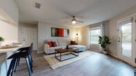This contemporary apartment showcases an open floor plan with a seamless flow between living spaces. The living room features a comfortable sectional sofa, modern artwork, and a ceiling fan for comfort. The adjacent kitchen boasts white cabinetry, stainless steel appliances, and a trendy backsplash. The bedroom is a highlight, with a king-size bed adorned in vibrant yellow accents, sliding glass doors leading to a private outdoor space, and ample natural light. The bathroom offers a clean, modern aesthetic with a shower/tub combination. Throughout the apartment, wood-look flooring and neutral tones create a cohesive, inviting atmosphere.