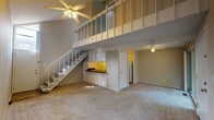 This two-story chalet-style residence offers a thoughtfully designed layout that makes excellent use of its space across two floors. The main level features an open living area with plush carpeting, soaring double-height ceilings, and a ceiling fan. A staircase with a white banister leads up to an open loft above, which overlooks the living space below and adds architectural character to the interior. The compact kitchen is efficiently laid out with white cabinetry, dark countertops, a stainless steel sink, and a yellow-toned backsplash accent. A tall window near the entryway draws in natural light, enhancing the airy feel of the main level. The upper floor hosts the bedroom, which features vaulted ceilings with exposed beams, a sliding glass door opening to a private balcony with outdoor views, and a mirrored closet. An additional room off a hallway provides flexible living space with its own closet and ceiling fan. The full bathroom is finished with a wide vanity, dark countertop, an integrated sink, and tile flooring. Throughout the home, neutral tones and carpet flooring create a warm, cohesive atmosphere suited for comfortable everyday living.