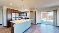 This well-appointed single-floor residence offers a thoughtfully designed layout that balances modern style with everyday functionality. The heart of the home is an open-concept kitchen, anchored by a large central island with a white quartz countertop, an integrated sink, and a dishwasher. Rich, dark-stained cabinetry lines the walls and provides generous storage, while a full suite of stainless steel appliances—including a refrigerator, range, and built-in microwave—delivers a polished, contemporary aesthetic. Pendant lighting over the island and recessed ceiling lights illuminate the space. A sliding glass door connects the kitchen and living area directly to an outdoor balcony, enhancing the sense of openness. Throughout the main living areas, durable light-toned hardwood-style flooring runs underfoot, while the bedrooms feature plush carpeting for added comfort. The spacious living area benefits from multiple large windows that frame outdoor greenery and allow abundant natural light to fill the room. The bedrooms are generously proportioned, each with ceiling fixtures and windows offering outdoor views. A dedicated laundry area houses a stacked washer and dryer unit for added convenience. The entry hallway includes an electrical panel, and the clean, neutral color palette throughout creates a bright and welcoming atmosphere.