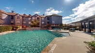This two-story residential community is defined by its impressive outdoor amenity area, centered around a generously sized swimming pool with crystal-clear turquoise water. The pool area is designed for both relaxation and socializing, featuring a broad concrete sundeck lined with teal-cushioned lounge chairs and a shallow wading ledge. Stone-accented steps and raised landscaped areas border the pool, adding a refined, layered aesthetic to the outdoor space.

A wood-framed pergola provides a shaded gathering spot alongside outdoor dining tables and chairs, ideal for al fresco entertaining. Mature palm trees and decorative plantings are positioned throughout the deck, enhancing the resort-like atmosphere. The pool is enclosed by ornamental iron fencing, ensuring both safety and a well-defined perimeter.

Adjacent to the pool is a substantial clubhouse building, distinguished by its blue-gray exterior, stone base accents, large grid-pane windows, and a prominent cupola atop the roofline. The structure appears to house interior amenity spaces and is directly accessible from the pool deck. The surrounding multi-story residential buildings feature a mix of stone and siding facades, pitched rooflines with dormers, and private balconies, all contributing to a cohesive architectural character throughout the community.
