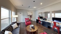 This single-floor business center is thoughtfully designed to balance professional functionality with a welcoming, stylish ambiance. The space features rich hardwood flooring throughout, crown molding details, and soft gray walls that create a calm, focused atmosphere.

Along one wall, a sleek built-in workstation provides multiple computer setups equipped with monitors and a printer, complemented by bold red industrial-style chairs and framed wall mirrors that add visual depth and a modern edge. Storage is integrated seamlessly beneath the desk surface, keeping the workspace organized and efficient.

The lounge area is furnished with a plush charcoal sofa, vibrant red accent chairs with decorative cushions, and a warm wooden coffee table — creating an inviting setting for informal meetings or relaxed breaks. Curated artwork adds personality throughout the space.

Natural light fills the room through multiple windows, while recessed ceiling lighting and floor lamps ensure consistent illumination at all hours. A glass-paned French door connects distinct areas of the center, maintaining an open feel while offering a sense of separation. The overall design strikes a balance between a productive work environment and a comfortable, design-forward gathering space.