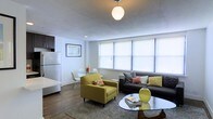 This well-appointed single-floor space showcases a thoughtfully designed open-concept layout that seamlessly blends the living, dining, and kitchen areas into one cohesive environment. The living area is anchored by a sleek dark sofa and a distinctive chartreuse accent chair with mid-century modern styling, complemented by a glass coffee table and a textured area rug that adds warmth to the hardwood flooring. A spherical pendant light serves as a focal point overhead, casting a soft glow throughout the space. A row of wide windows fitted with roller shades allows natural light to fill the room, creating a bright and airy ambiance. The compact dining area features a round table with clean-lined white chairs, positioned conveniently adjacent to the living space. The kitchen, visible from the main living area, includes dark upper cabinetry and a full-size refrigerator, maintaining a functional yet integrated presence. The textured ceiling adds a subtle architectural detail, while the neutral wall tones provide a versatile backdrop that enhances the vibrancy of the colorful furnishings. The overall aesthetic is modern, curated, and inviting.