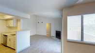 This single-floor space offers a clean, functional layout designed for comfortable everyday living. The main living area is bathed in natural light streaming through a large window fitted with horizontal blinds, highlighting the light-toned hardwood flooring that runs throughout. A textured popcorn ceiling adds a classic touch overhead, while the neutral beige and white wall palette keeps the space feeling open and airy. A built-in fireplace serves as a focal point in the living room, providing both a practical heating element and a warm aesthetic anchor to the space. A partial wall with a raised counter suggests a kitchen pass-through or breakfast bar, hinting at a connected kitchen area just beyond. A doorway visible in the background leads to additional rooms, indicating a thoughtfully segmented floor plan that balances open communal areas with more private spaces. The overall ambiance is understated and versatile, offering a neutral canvas well-suited for a variety of interior styles. The space presents a straightforward, move-in-ready environment with essential features that prioritize both comfort and practicality.