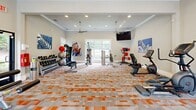 Located at 5250 Highway 138 in Union City, Georgia, this fitness center offers a thoughtfully designed exercise room that provides ample space for a comprehensive workout experience. The room features elegant crown molding, recessed lighting, and dual ceiling fans that keep the environment comfortable during intense sessions. Large windows along the walls flood the space with natural light while offering pleasant views of the surrounding greenery outdoors.

The exercise room is outfitted with a diverse array of equipment to support varied fitness routines. Cardio options include multiple treadmills, a recumbent bike, and a stair-stepping machine, while strength training is supported by a cable/functional trainer and a dedicated weight bench. Free weight offerings include a full dumbbell rack, kettlebells, medicine balls, and stability balls stored on a TAG-branded rack. Wall-mounted flat-screen televisions provide entertainment during workouts, and vibrant fitness-themed artwork decorates the walls, adding energy to the space.

The distinctive patterned carpet in orange, gray, and white ties the room together visually and provides a durable, cushioned surface underfoot. A glass entry door with an illuminated exit sign ensures safe and easy access. Overall, this fitness center delivers a bright, motivating, and fully equipped environment for residents seeking an active lifestyle.