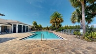 Located in Tavares, Florida, this outdoor community amenity area offers a quintessential Florida resort experience. The centerpiece of the space is a generously sized swimming pool with crystal-clear turquoise water, complemented by a separate spa and a dedicated wading or leisure pool area. The pools are surrounded by an expansive paver deck crafted from multi-toned stone pavers, providing ample room for lounging and socializing under the Florida sun.

Numerous lounge chairs and seating areas are thoughtfully arranged around the pool deck, inviting relaxation in the open air. A covered gazebo on one end of the deck provides a shaded retreat, while tall, swaying palm trees frame the perimeter, enhancing the tropical ambiance throughout.

An adjacent clubhouse-style building with a covered colonnade and large windows opens directly onto the pool deck, seamlessly connecting indoor and outdoor spaces. The facility is enclosed by a decorative fence, ensuring privacy while still allowing sweeping views of the surrounding lush greenery and neighboring community. Under the bright blue Florida sky, this community pool area captures the essence of warm-weather outdoor living, offering an inviting and well-appointed gathering space for residents.