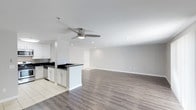 This single-story space showcases an open-concept layout that seamlessly connects the kitchen and living areas, creating a bright, airy atmosphere. The kitchen features white shaker-style cabinetry paired with dark countertops, offering a clean contrast that adds visual interest. A peninsula extends from the kitchen, providing additional workspace and a natural transition into the main living area. Stainless steel appliances and light tile flooring complete the kitchen zone. The expansive living area is finished with warm wood-look laminate flooring, adding texture and continuity throughout the space. Recessed lighting is installed throughout the ceiling, ensuring even, ambient illumination, while a ceiling fan with integrated lighting adds both comfort and a functional focal point. Neutral gray walls provide a versatile backdrop suitable for a variety of décor styles. The layout is notably open, offering generous room for flexible furniture arrangements. The overall design strikes a balance between modern and practical, making it a functional and visually cohesive living environment.