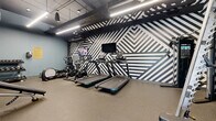 Located at 4757 Hiawatha Avenue in Minneapolis, MN, this fitness center is a well-appointed, single-level exercise facility designed to deliver a full-featured workout experience within an efficiently organized footprint. The space immediately draws attention with its striking black-and-white geometric accent wall, featuring bold diagonal stripe patterns that lend a modern, energetic aesthetic to the room.

The flooring throughout consists of durable rubber gym matting, providing safety and support for a wide range of exercises. Overhead, an exposed ceiling with black-painted ductwork and linear LED lighting creates an industrial-modern atmosphere while ensuring the space is brightly and evenly lit.

Cardio equipment is well-represented, with multiple treadmills, stationary spin bikes, an elliptical trainer, and a rowing machine positioned along the accent wall. Mounted flat-screen televisions are conveniently placed above the cardio stations. On the strength training side, the facility includes a power rack with a barbell, an adjustable weight bench, and multiple dumbbell racks stocked with a range of weights. A large wall mirror amplifies the sense of space and allows users to monitor their form.

Soft blue-gray walls on the perimeter complement the bold accent wall, and a water fountain and sanitation station are accessible near the entrance, which features a glass door allowing natural light to filter in.