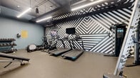 Located at 4757 Hiawatha Avenue in Minneapolis, MN, this fitness center is a well-appointed, single-level exercise facility designed to deliver a full-featured workout experience within an efficiently organized footprint. The space immediately draws attention with its striking black-and-white geometric accent wall, featuring bold diagonal stripe patterns that lend a modern, energetic aesthetic to the room.

The flooring throughout consists of durable rubber gym matting, providing safety and support for a wide range of exercises. Overhead, an exposed ceiling with black-painted ductwork and linear LED lighting creates an industrial-modern atmosphere while ensuring the space is brightly and evenly lit.

Cardio equipment is well-represented, with multiple treadmills, stationary spin bikes, an elliptical trainer, and a rowing machine positioned along the accent wall. Mounted flat-screen televisions are conveniently placed above the cardio stations. On the strength training side, the facility includes a power rack with a barbell, an adjustable weight bench, and multiple dumbbell racks stocked with a range of weights. A large wall mirror amplifies the sense of space and allows users to monitor their form.

Soft blue-gray walls on the perimeter complement the bold accent wall, and a water fountain and sanitation station are accessible near the entrance, which features a glass door allowing natural light to filter in.