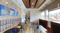 This single-floor space is designed as a sophisticated lounge or café-style gathering area, blending contemporary design elements with warm, inviting textures. The interior immediately draws attention to its striking ceiling detail, where rich wood slat panels are inset within a recessed tray ceiling, complemented by recessed lighting that provides a warm, ambient glow throughout the space.

The flooring features polished large-format tile that reflects light and enhances the sense of openness. One wall showcases exposed natural stone or brick cladding, adding a rustic, urban character that contrasts beautifully with the clean white walls. Contemporary artwork adorns the walls, contributing to the space's modern aesthetic.

Seating is thoughtfully varied, incorporating a built-in banquette with striped upholstery along one wall, paired with sleek café-style bistro tables and industrial-inspired chairs. A window bar counter along the opposite wall offers additional seating with views of the surrounding urban landscape, as large windows allow generous natural light to flood the interior. A decorative starburst wall clock serves as a focal point, tying together the modern-eclectic design sensibility throughout.
