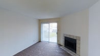 This single-floor space presents a clean, straightforward layout centered around a comfortable living area. The room is finished with wall-to-wall carpeting in a neutral tone, providing a warm foundation throughout. A standout feature is the fireplace, framed by a classic tile surround in a beige and tan palette, adding a functional and visual focal point to the space.

Natural light fills the room through a full-length sliding glass door fitted with vertical blinds, offering both privacy and easy access to the outdoors. The door provides a view of a green exterior area, connecting the interior to the outside environment. The walls are painted in a light, neutral white, contributing to a bright and open feel.

The ceiling is smooth and consistently level, complementing the room's clean lines. The overall ambiance is understated and practical, offering a blank canvas well-suited for a variety of furnishing styles. The combination of the fireplace, natural light, and neutral finishes makes this a functional and inviting living space.