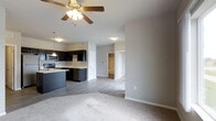 This single-floor space showcases a thoughtfully designed open-concept layout that seamlessly connects the living and kitchen areas. The living space features plush carpeting and a ceiling fan with integrated lighting, creating a comfortable and well-ventilated environment. The transition to the kitchen is marked by a shift to smooth hardwood-style flooring, lending a clean and modern aesthetic throughout.

The kitchen is outfitted with dark espresso cabinetry that provides a striking contrast against the neutral gray walls. Stainless steel appliances, including a range and microwave, are integrated alongside a kitchen island that offers additional prep space and storage. A deep basin sink is positioned beneath pendant lighting, adding a contemporary touch to the culinary space.

The neutral gray walls and light-toned ceilings create a cohesive, understated palette that allows natural light to brighten the interior. Doorways visible from the main living area lead to additional rooms, including what appears to be a bedroom with a walk-in closet, suggesting a well-organized and functional floor plan. White trim and clean baseboards throughout add a polished, finished look to the overall space.