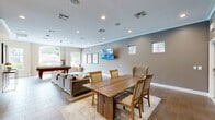 This single-floor community amenity space offers a thoughtfully designed open-concept layout that seamlessly integrates multiple functional areas. The main gathering area is expansive and bright, featuring large windows and a glass door that flood the space with natural light and provide views of the surrounding outdoor greenery. Crown molding, recessed lighting, and tile flooring that mimics warm wood run throughout, lending a cohesive and polished aesthetic.

The lounge area is anchored by a generous sectional sofa, ottomans, and a wall-mounted flat-screen television, creating an inviting setting for relaxation. Adjacent to this is a game room zone centered around a full-size pool table beneath a striking modern chandelier, with additional seating and artwork completing the area. A dedicated dining space features a solid wood dining table surrounded by upholstered chairs, ideal for group gatherings.

The kitchen is sleek and contemporary, outfitted with taupe cabinetry, open floating shelves, white subway tile backsplash, light quartz countertops, a stainless steel refrigerator with ice dispenser, and a gold-tone faucet. A hallway connects the main areas and provides access to a restroom. Together, these spaces form a versatile and well-equipped communal environment.