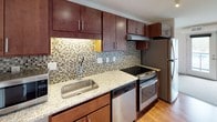 This single-floor unit offers a thoughtfully designed layout that balances modern finishes with practical functionality. The kitchen is a standout feature, showcasing rich dark-wood cabinetry that extends to the ceiling for ample storage, complemented by granite countertops, a mosaic tile backsplash, and a full suite of stainless steel appliances including a gas range, dishwasher, and refrigerator. Under-cabinet lighting adds warmth and enhances the workspace. Hardwood flooring runs through the kitchen, transitioning seamlessly into a carpeted living area that provides a comfortable and cozy atmosphere. A window in the living space allows natural light to filter in while offering views of the surrounding exterior. The bedroom is quietly understated, featuring neutral walls, plush carpet, and a window with an outdoor view, creating a calm retreat. The bathroom is generously sized and accessibility-conscious, featuring a rich wood vanity with granite countertop, a large mirror with overhead lighting, and safety grab bars. The overall design throughout the unit emphasizes clean lines, neutral tones, and quality materials, resulting in a cohesive and livable space.