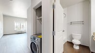 This thoughtfully designed studio unit makes efficient use of its single-floor layout, offering a clean, modern aesthetic throughout. The open-plan living and kitchen area is anchored by a well-appointed kitchen featuring light blue lower cabinetry, crisp white upper cabinets, a stainless steel refrigerator, a built-in microwave, a cooktop, and a single-basin sink — all set against a bright white countertop and backsplash. A wall-mounted fold-down counter provides a practical dining or workspace solution. Light-toned laminate wood flooring runs throughout the main living area, lending warmth and continuity to the space. The living area is naturally lit by a large multi-pane window offering outdoor views, and includes a wall-mounted PTAC heating and cooling unit alongside a tall built-in wardrobe for additional storage. The bathroom is finished with large-format dark tile flooring, a granite-topped vanity, and a shower, conveying a grounded, clean design. Tucked between the bathroom and living area is a dedicated laundry closet housing a front-loading washer, adding everyday convenience. Recessed lighting and neutral wall tones throughout create a bright, airy ambiance well-suited to modern urban living.