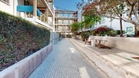 Located at 2020 4th St in Berkeley, California, this expansive outdoor courtyard serves as a well-designed communal gathering space set between contemporary multi-story residential buildings. The courtyard is defined by a long, paver-lined walkway that runs through its center, flanked by a series of raised concrete planter beds filled with lush greenery, ornamental grasses, shrubs, and mature trees that provide natural shade and visual interest throughout the space. On one side, tiered concrete steps create a terraced seating area with Adirondack-style wooden chairs, offering a relaxed setting for outdoor enjoyment. String lights are strung overhead, adding a warm, ambient atmosphere suited for evening gatherings. A dedicated outdoor kitchen area with a built-in BBQ grill and counter space is conveniently positioned at one end of the courtyard, making the space well-suited for outdoor entertaining. Raised garden planters with a variety of plantings line portions of the perimeter, adding texture and a verdant character to the hardscape. The surrounding buildings feature balconies with cable railings and wood accents that overlook the courtyard, while a pergola structure at the far end frames views and reinforces the architectural cohesion of the space. The courtyard benefits from abundant natural sunlight, creating an inviting, open-air environment that balances functional amenities with thoughtful landscaping.