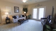 This single-floor space offers a welcoming and well-appointed living environment designed for comfortable everyday living. The living room serves as the centerpiece of the home, featuring plush wall-to-wall carpeting in a soft neutral tone that adds warmth underfoot. A ceiling fan with a light fixture is centrally mounted, providing both air circulation and ambient lighting. The room is anchored by a light-colored sofa accented with decorative navy and patterned throw pillows, paired with a dark wood coffee table that adds a striking contrast to the lighter surroundings. A tall, lush potted plant in the corner brings a touch of natural greenery to the interior. The walls are finished in a warm beige tone, complemented by decorative wall art and ornamental metalwork above the doorway, adding character and personality to the space. A prominent set of white-framed French doors fills the room with natural light, offering views to the exterior and creating an airy, open atmosphere. The overall ambiance is cozy yet refined, with a harmonious blend of neutral tones and tasteful décor that makes the space feel inviting and move-in ready.