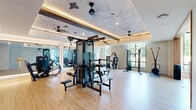 Located at 412 Summer St in Abington, MA, this single-floor fitness facility offers a thoughtfully designed exercise environment spread across two dedicated exercise rooms. The main gym area impresses with soaring double-height ceilings adorned with distinctive wave-pattern wall art and warm recessed cove lighting. Light wood flooring runs throughout, complementing the bright, airy atmosphere created by floor-to-ceiling windows that frame views of the surrounding greenery. This primary space is comprehensively equipped with cardio machines including treadmills and stationary bikes, a multi-station cable machine, strength equipment, a punching bag, and a squat rack, catering to a wide range of workout styles. Large mirrors line the walls, visually expanding the space further. Ceiling fans maintain airflow throughout. The second exercise room serves as a dedicated studio space, featuring sage green accent walls, mirrored walls, and large windows that flood the room with natural light and outdoor views. This room is outfitted with Pilates reformers, balance tools such as a BOSU ball, resistance accessories, and a wall-mounted storage rack for additional equipment. Together, the two rooms provide a comprehensive and well-appointed fitness experience.