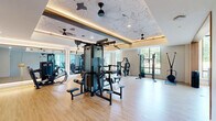 Located at 412 Summer St in Abington, MA, this single-floor fitness facility offers a thoughtfully designed exercise environment spread across two dedicated exercise rooms. The main gym area impresses with soaring double-height ceilings adorned with distinctive wave-pattern wall art and warm recessed cove lighting. Light wood flooring runs throughout, complementing the bright, airy atmosphere created by floor-to-ceiling windows that frame views of the surrounding greenery. This primary space is comprehensively equipped with cardio machines including treadmills and stationary bikes, a multi-station cable machine, strength equipment, a punching bag, and a squat rack, catering to a wide range of workout styles. Large mirrors line the walls, visually expanding the space further. Ceiling fans maintain airflow throughout. The second exercise room serves as a dedicated studio space, featuring sage green accent walls, mirrored walls, and large windows that flood the room with natural light and outdoor views. This room is outfitted with Pilates reformers, balance tools such as a BOSU ball, resistance accessories, and a wall-mounted storage rack for additional equipment. Together, the two rooms provide a comprehensive and well-appointed fitness experience.