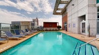 Located at 848 Spring St NW in Atlanta's Midtown neighborhood, this rooftop amenity level offers an impressive outdoor retreat with sweeping views of the Atlanta skyline, including notable landmarks such as Georgia Tech's campus. At the heart of the space is a generously sized swimming pool with crystal-clear turquoise water, framed by a broad concrete pool deck lined with blue lounge chairs ideal for sunbathing. The surrounding architecture features a striking combination of exposed concrete, brick, and large floor-to-ceiling glass panels, lending the space a modern, industrial aesthetic. A covered pergola area with warm wood detailing and string lights provides a shaded lounge zone furnished with contemporary tables and chairs set on artificial turf, perfect for casual gatherings. Manicured topiaries and landscaped greenery add natural accents throughout the deck. Glass railings along the perimeter ensure unobstructed panoramic views of the cityscape on all sides. The space is well-suited for both relaxation and socializing, offering a dynamic urban outdoor environment elevated above the city.