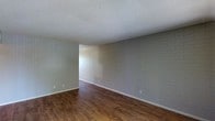This single-floor space presents a straightforward and functional design, highlighted by several distinctive architectural details. The most notable feature is the painted brick wall, finished in a light gray tone that adds texture and character to the interior without overwhelming the overall aesthetic. This accent wall runs the length of the main living area, providing a subtle industrial touch that contrasts pleasantly with the otherwise neutral palette.

The flooring throughout consists of wood-look planks in a warm, medium-brown finish, lending a sense of warmth and continuity to the space. The planks are laid uniformly, contributing to a clean, cohesive look underfoot. White baseboards provide a crisp border between the floor and walls.

Overhead, a textured popcorn ceiling spans the room, a characteristic detail that reflects the building's construction era. The walls are painted in a soft, muted gray-green tone, complementing both the brick feature wall and the flooring.

An open passage visible in the background suggests a connected layout, allowing for a natural flow between areas. The space is clean, freshly updated, and move-in ready, offering a neutral foundation suited for a variety of interior styles.
