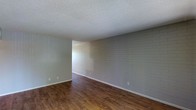 This single-floor space presents a straightforward and functional design, highlighted by several distinctive architectural details. The most notable feature is the painted brick wall, finished in a light gray tone that adds texture and character to the interior without overwhelming the overall aesthetic. This accent wall runs the length of the main living area, providing a subtle industrial touch that contrasts pleasantly with the otherwise neutral palette.

The flooring throughout consists of wood-look planks in a warm, medium-brown finish, lending a sense of warmth and continuity to the space. The planks are laid uniformly, contributing to a clean, cohesive look underfoot. White baseboards provide a crisp border between the floor and walls.

Overhead, a textured popcorn ceiling spans the room, a characteristic detail that reflects the building's construction era. The walls are painted in a soft, muted gray-green tone, complementing both the brick feature wall and the flooring.

An open passage visible in the background suggests a connected layout, allowing for a natural flow between areas. The space is clean, freshly updated, and move-in ready, offering a neutral foundation suited for a variety of interior styles.