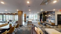 This expansive single-floor amenity space showcases a modern-industrial design that blends exposed brick columns, rustic wood beam accents, and polished concrete flooring with plush carpeted zones. Large floor-to-ceiling windows along the perimeter flood the interior with natural light and provide views of the surrounding exterior, enhancing the open, airy atmosphere throughout.

The space is organized into several distinct activity and lounge areas. A standout feature is the indoor putting green, set against the window wall and complemented by seating nearby. Adjacent to it, a shuffleboard table and arcade games offer additional entertainment options. Exposed brick columns anchor the layout, serving as architectural focal points that divide zones while maintaining visual continuity.

A dedicated lounge area features a deep sectional sofa, ottomans, and a wall-mounted television, alongside a bold teal wainscoted accent wall decorated with artistic wall hangings and a large board game display. A dining and collaboration zone includes a long communal table with upholstered chairs, pendant lighting overhead, and decorative wall art. Recessed lighting throughout ensures consistent illumination, while the varied textures of brick, wood, concrete, and soft furnishings contribute to a warm yet contemporary ambiance.