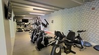 This fitness center offers a comprehensive workout environment designed to accommodate a range of exercise needs. The space is outfitted with an impressive selection of cardio equipment, including elliptical trainers, upright and recumbent stationary bikes, and treadmills, all arranged to allow comfortable use and easy movement throughout the room. A dedicated strength training area is also visible, featuring a cable machine and additional equipment to support a full-body workout routine. The flooring incorporates a durable, cushioned surface suited for high-traffic gym use, with a red mat section designating a functional training zone. Overhead, long fluorescent light fixtures are suspended from exposed ceiling beams, providing bright, even illumination throughout the space. A wall-mounted flat-screen television adds an entertainment element for those using the cardio machines. The standout design feature is a large, bold geometric-patterned accent wall in a monochromatic palette, lending the space a modern and visually dynamic character. The overall ambiance is clean, functional, and purposefully designed, striking a balance between an inviting aesthetic and a practical, well-organized fitness facility.